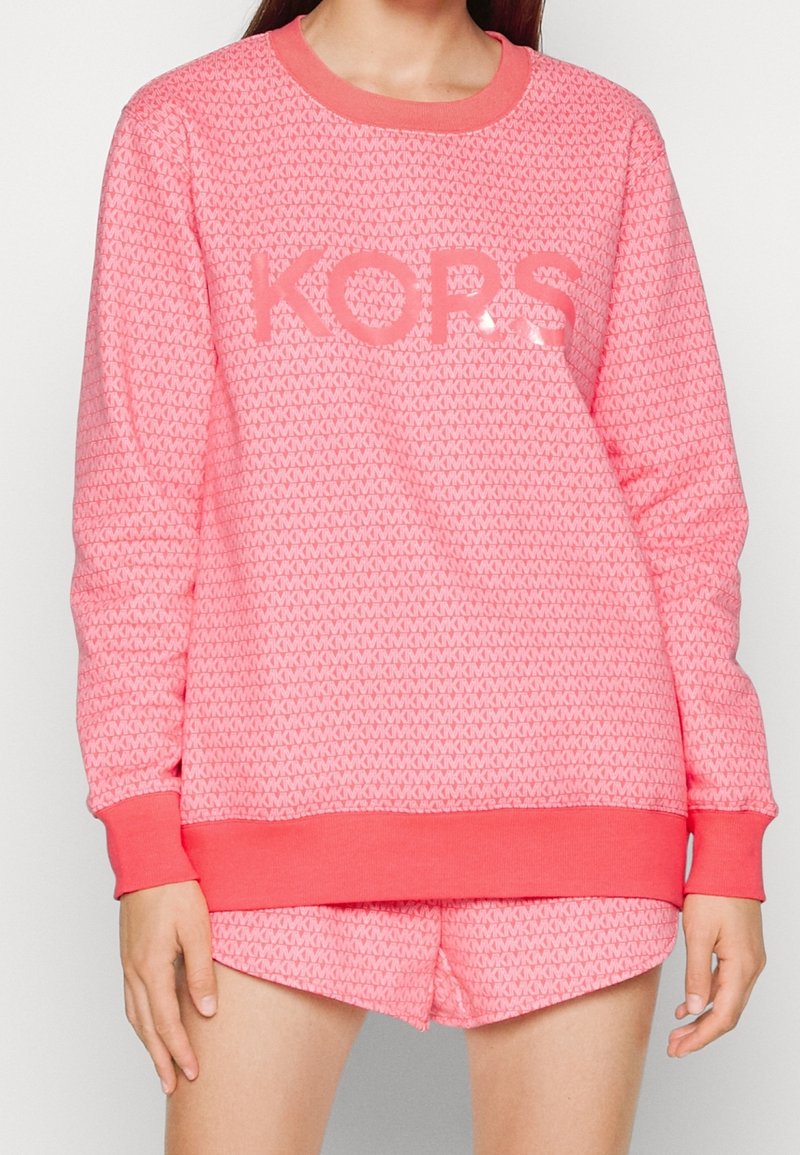 Femme portant un sweat-shirt rose à manches longues et un short assorti avec un motif répétitif "MK" et le logo "KORS" sur le devant.