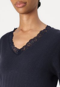 Pull en maille bleu marine avec un décolleté en V, orné d'une délicate bordure en dentelle le long de l'encolure, mettant en valeur une texture douce et une coupe ajustée.