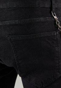 Melnas denim bikses, kurās redzams tuvplāns aizmugurējai kabatai ar redzamām dūrienu šuvēm un metāla ķēdes akcentu. Audums izskatās nedaudz nodilis.