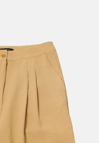 Béžové kalhoty s vysokým pasem, předními záhyby, bočními kapsami, zapínáním na knoflík a hladkým texturovaným materiálem na bílém pozadí.