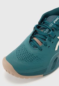Tyrkysová sportovní bota s texturovaným svrškem, síťovinovými akcenty a béžovou gumovou podrážkou. Obsahuje šněrování a prvky brandingu. Název modelu je viditelný.