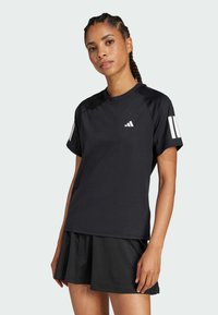 Tricou sport negru cu mâneci scurte, având un mic logo alb și trei dungi albe pe mâneca dreaptă, purtat cu o fustă plisată neagră.