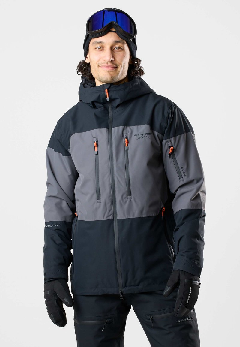 Homme portant une veste d'hiver noire et grise, des gants noirs et un masque de ski bleu sur le front, souriant devant un fond uni.