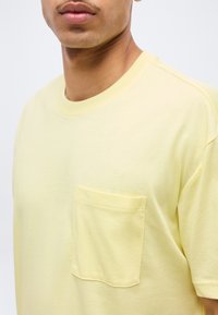 Κοντινή λήψη ενός ατόμου που φοράει ανοιχτό κίτρινο κοντομάνικο μπλουζάκι με τσέπη στο στήθος, εμφανίζοντας μέρος του λαιμού και των ώμων.