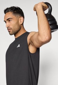 Egy férfi a jobb kezével fekete kettlebellt emel, ujjatlan fekete edzőpólót visel, amelyen fehér logó található, összpontosító arckifejezéssel, világos háttér előtt.