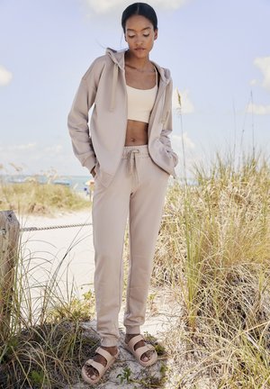 Beige Kapuzenpullover und passende Jogginghose, gefertigt aus weichem Material. Bemerkenswerte Details sind Kordeln, gerippte Akzente und geschnallte Sandalen.