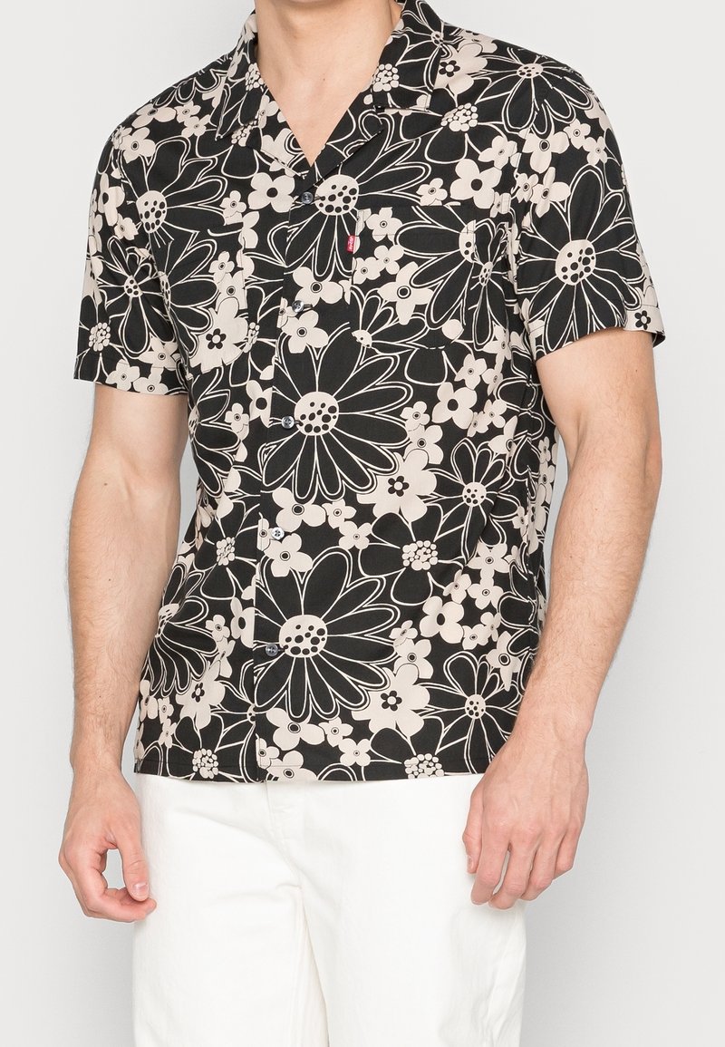 Camicia a maniche corte nera con motivo floreale bianco. Presenta una chiusura con bottoni davanti e una tasca sul petto. Realizzata in tessuto leggero.