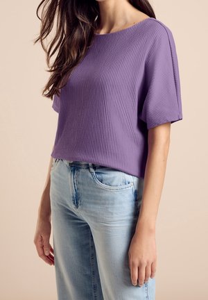 Frau trägt ein lila, geripptes Kurzarmshirt, kombiniert mit hellblauen, hoch taillierten Jeans vor einem beigen Hintergrund.