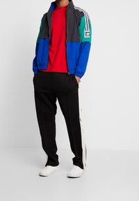 Färgglad jacka i blått, grönt och grått med vita ränder; röd skjorta; svarta byxor med vita sidränder; vita sneakers.