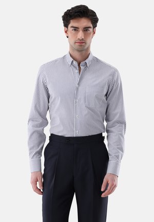 Young man wearing a fitted white dress shirt with navy check pattern and dark pleated trousers, standing against a white background.