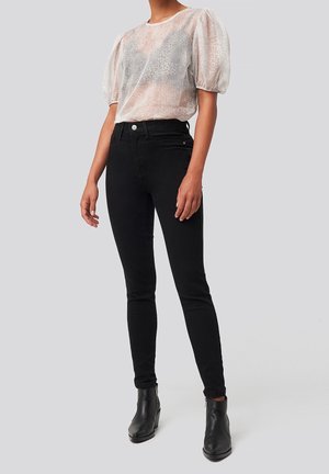 Haut transparent à manches bouffantes avec un imprimé animalier rose clair et gris, associé à un jean skinny noir et des bottines, mettant en valeur une silhouette ajustée.