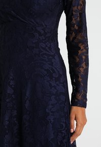 Granatowa koronkowa sukienka z misternymi kwiatowymi wzorami, długimi przezroczystymi rękawami i fakturowanym materiałem. Dopasowana góra z rozkloszowanym dołem.