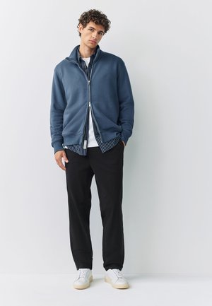 Giovane uomo con capelli ricci che indossa una giacca blu con zip sopra una camicia a righe, pantaloni neri e sneaker bianche, in piedi davanti a uno sfondo neutro.