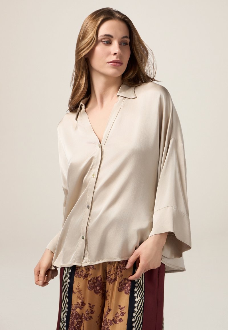 Blusa di seta beige chiaro con colletto, design morbido e maniche a tre quarti. Presenta bottoni discreti e una texture liscia.