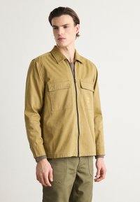 Svetlo rjav zip-up jopič iz trpežnega blaga, z dvema žepoma na prsih, ovratnikom in rebrastimi pletenimi manšetami. Obučen čez strukturirano majico.