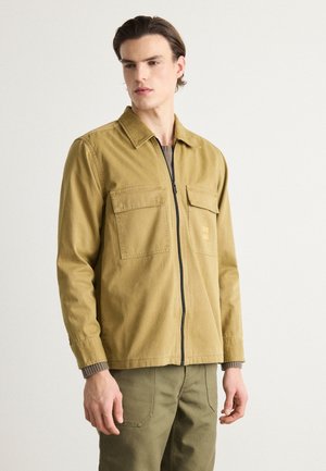 Veste zippée marron clair en tissu durable, dotée de deux poches poitrine, d'un col et de poignets en tricot côtelé. Portée par-dessus un top texturé.