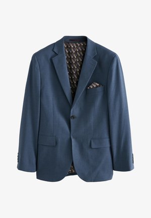 Mężczyzna w dopasowanym ciemnoniebieskim garniturze stoi tyłem do widza na jednolitym beżowym tle.