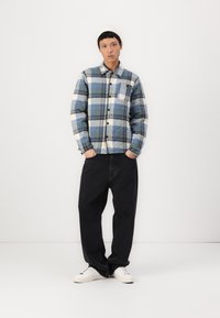 Quilted plaid shirt in blue, cream, and black with button closure and chest pocket, paired with loose black trousers and white sneakers.