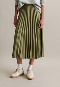 Person in einem olivgrünen, gefalteten Midirock, hellgrauem Pullover, weißen Socken und beigen Sneakern vor einem einfarbigen Hintergrund.