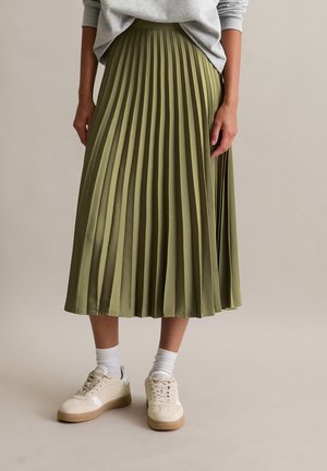 Person wearing olive green pleated midi skirt, light grey sweater, white socks, and beige sneakers against plain background.
