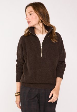 Maglione marrone con zip e collo alto, polsini a coste e lavorazione a maglia strutturata. Abbinato a pantaloni a quadri, che mostrano una vestibilità rilassata.