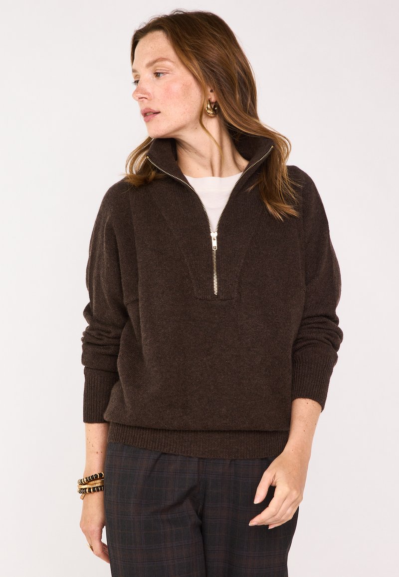 Pull en zip marron avec col montant, poignets côtelés et tricot texturé. Associe à un pantalon à carreaux, mettant en avant une coupe décontractée.