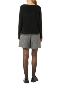 Schwarzer Rippstrick-Pullover mit einem silberfarbenen Saum, kombiniert mit grauen Hahnentritts-Shorts. Das Model trägt schwarze Strumpfhosen und Schuhe mit dicken Sohlen.