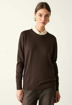 Kvinde iført en mørkebrun langærmet sweater over en lysebeige skjorte med krave og mørke bukser, stående mod en ensfarvet lys baggrund.