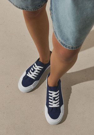 Jambes portant des baskets en toile bleue avec des lacets blancs et des bouts en caoutchouc blanc, debout sur un sol clair avec un short en jean.