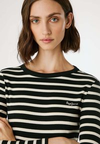 Melna un krēmkrāsas svītrainā krekla ar garām piedurknēm, rievotu struktūru, apaļu kakla izgriezumu, kas rotāts ar mazu "Pepe Jeans" logo kreisajā krūtīs.
