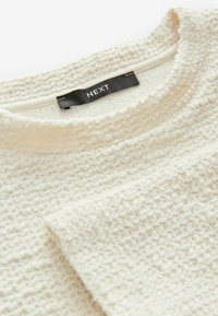 Pull texturé couleur crème avec un motif tissé, doté d'un col rond et d'une étiquette "NEXT" près du col.