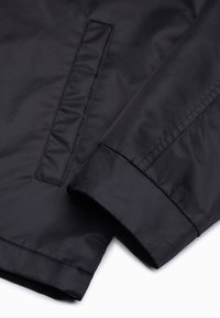 Manche de veste noire et détail de poche avec couture visible sur tissu lisse sur fond blanc.