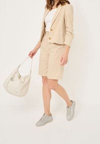 Femme portant un blazer beige et un short avec un haut blanc, portant un sac à main blanc et des baskets gris clair, marchant à l'intérieur.