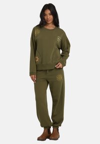 Felpa verde oliva e pantaloni abbinati, entrambi realizzati in tessuto morbido. Presentano motivi dorati a forma di sole e hanno una vestibilità rilassata, con polsini alle caviglie.