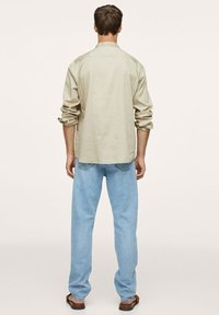 Jasnobeżowa koszula z guzikami, z podwiniętymi rękawami, w połączeniu z jasnoniebieskimi dżinsami i brązowymi sandałami, widok z tyłu.