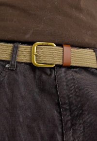 Cinturón tejido color caqui con hebilla de metal dorado y acento de cuero marrón, usado sobre pantalones de pana oscuros. Textura y contraste de color distintivos.