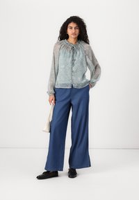 Himmelbluse mit Muster und Kordelzug-Ausschnitt, kombiniert mit weit geschnittenen dunklen Jeans und schwarzen Schuhen; das Model hält eine kleine weiße Tasche.