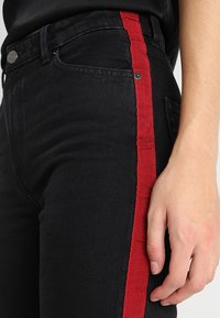 Černé džíny z denimového materiálu s výrazným červeným bočním pruhem. Tkanina má texturovaný povrch, s kovovými prvky a viditelnými detaily šití.