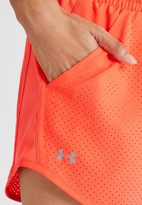 Jasnopomarańczowe szorty sportowe z perforowaną strukturą, elastycznym pasem i boczną kieszenią. Zawierają małe szare logo blisko dolnego brzegu.