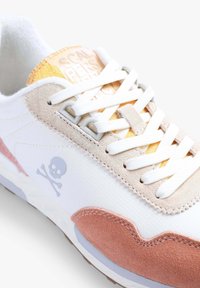 Sneaker caratterizzato da materiale in canvas bianco, accenti in suede beige e rosa, dettagli gialli sulla linguetta e un logo con teschio e ossa incrociate.