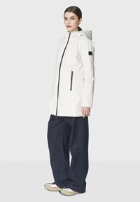 Nainen, jolla on yllään valkoinen huppari takki, jossa on mustat vetoketjulla varustetut taskut, tummat leveälahkeiset farkut ja valkoiset lenkkarit punaisilla yksityiskohdilla, seisoo sivuttain.