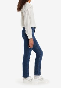 Femme debout de profil portant une chemise blanche rayée, un jean bleu, des chaussettes blanches et des baskets blanches sur un fond uni.