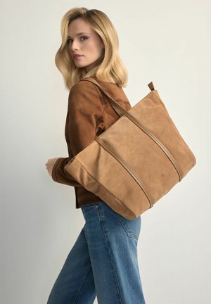 Blondynka w brązowej kurtce i niebieskich dżinsach niesie dużą jasnobrązową torbę z zamszu na ramieniu, patrząc za siebie.