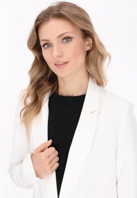 Bílý blazer s přiléhavým střihem, hladkou texturou a klopovým límcem, nošený přes černý top, s vlnitými, světle hnědými vlasy rámujícími obličej.