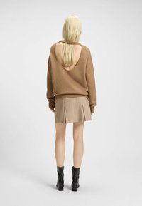Pull en maille marron avec une large découpe dans le dos, associé à une jupe plissée beige. Des bottines noires à talons épais complètent la tenue.