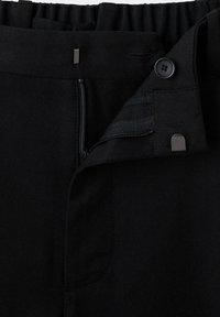 Ceinture de pantalon noire avec fermeture éclair partiellement ouverte, bouton noir, dos élastique et attaches métalliques à crochets et barres.