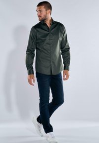 Mann in einem dunkelgrünen Hemd mit Knöpfen, dunkelbblauen Jeans und weißen Turnschuhen, der mit einem gebeugten Bein steht und zur Seite schaut.