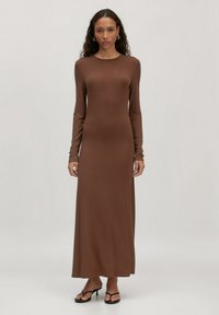 Frau mit langen lockigen Haaren, die ein enges braunes Maxikleid mit langen Ärmeln und schwarze Absatzsandalen trägt, steht vor einem schlichten Hintergrund.