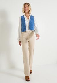Chaleco acolchado azul sobre una blusa blanca, combinado con pantalones de campana beiges y sandalias de plataforma marrones. Diseño sencillo con un aspecto texturizado.