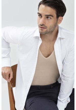 Weiße mit Knöpfen versehene Bluse über einem beigefarbenen Top mit glatter Textur, vor einem hellen Hintergrund. Modell sitzt auf einem Holzstuhl.